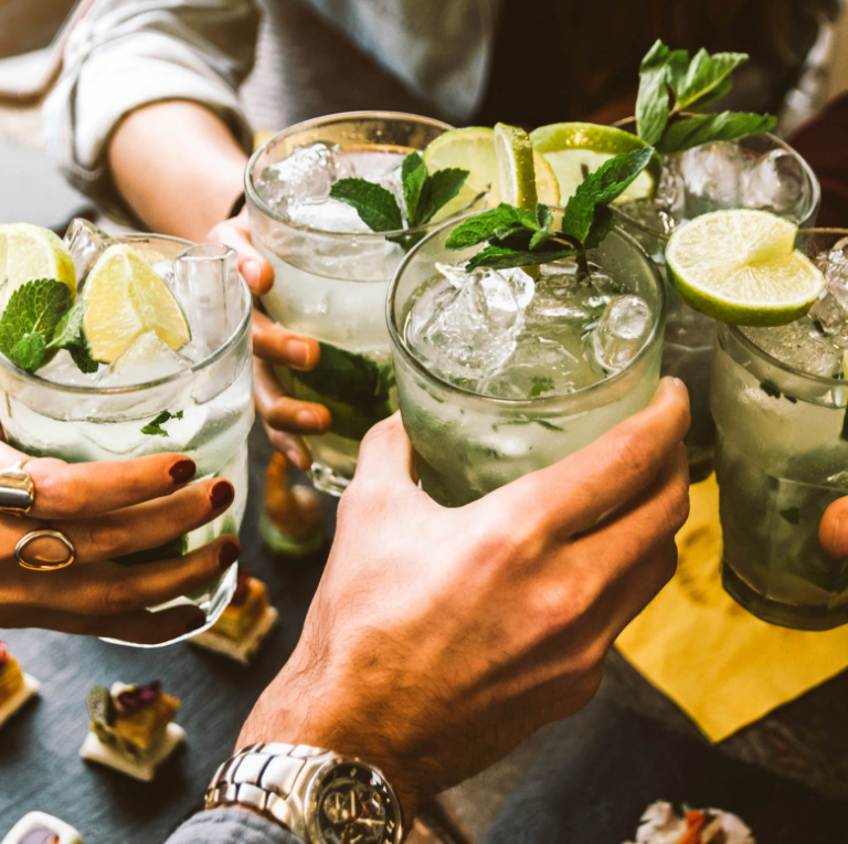 Friends clink cocktails with lime wedges and mint at a table outdoors.