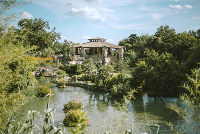 A wooden pavilion with a tiered roof surrounded by green trees and shrubs beside a pond under a partly cloudy sky.
