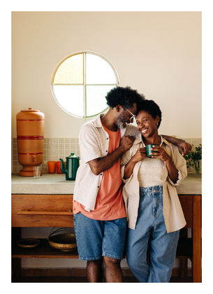 Two people stand close together in a kitchen beside a round wall mirror, a wooden cabinet, and potted plants.