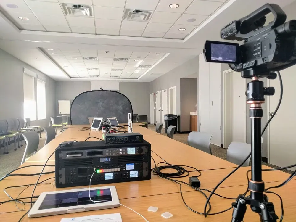 Conference room with video camera on tripod, audio equipment, and cables set up for a recording session