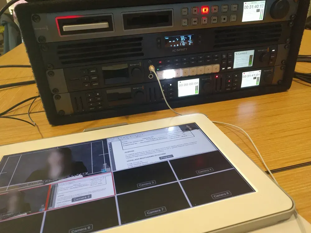 Tablet on a wooden table showing a control interface, with stacked AV equipment and lit displays behind it