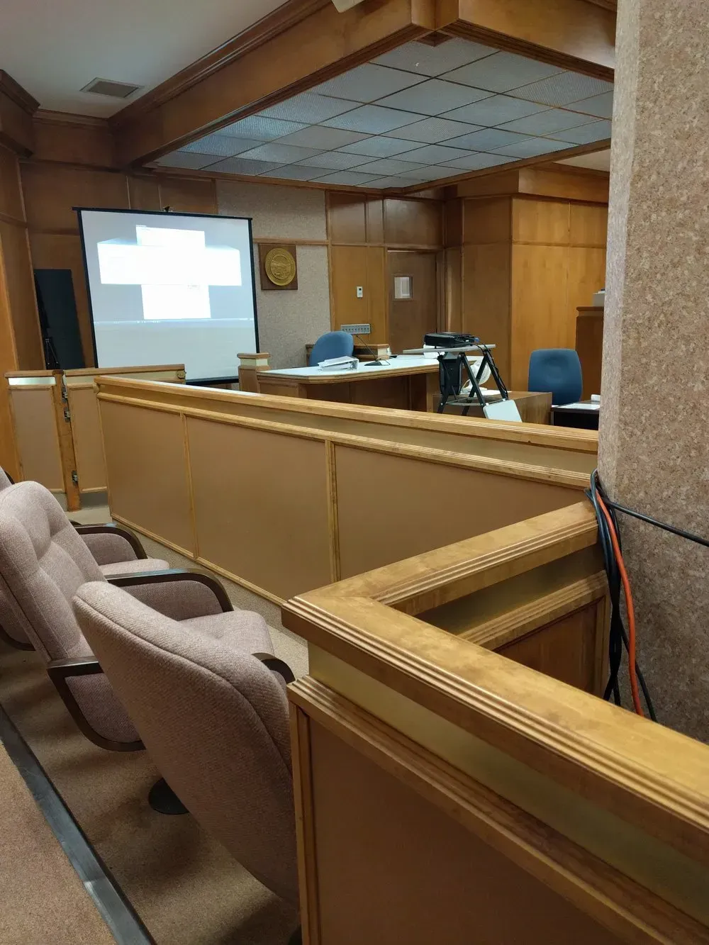 Empty courtroom with wood paneling, witness stand, counsel tables, and a projected screen at the front.