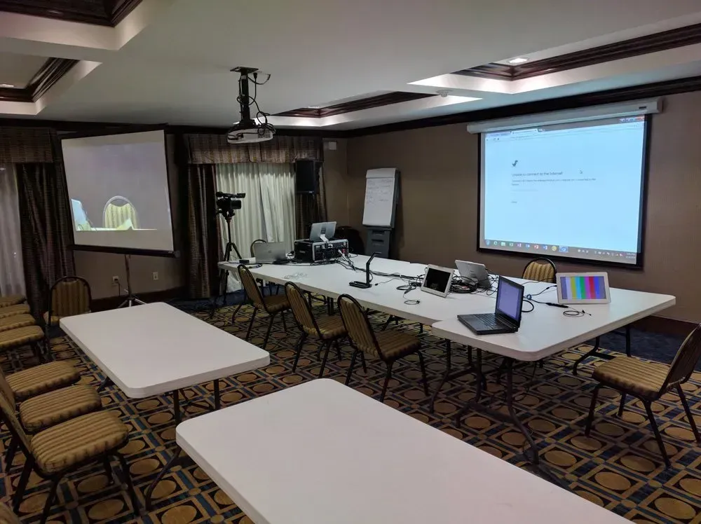 Conference room with two projection screens, tables, chairs, laptops, and a flip chart
