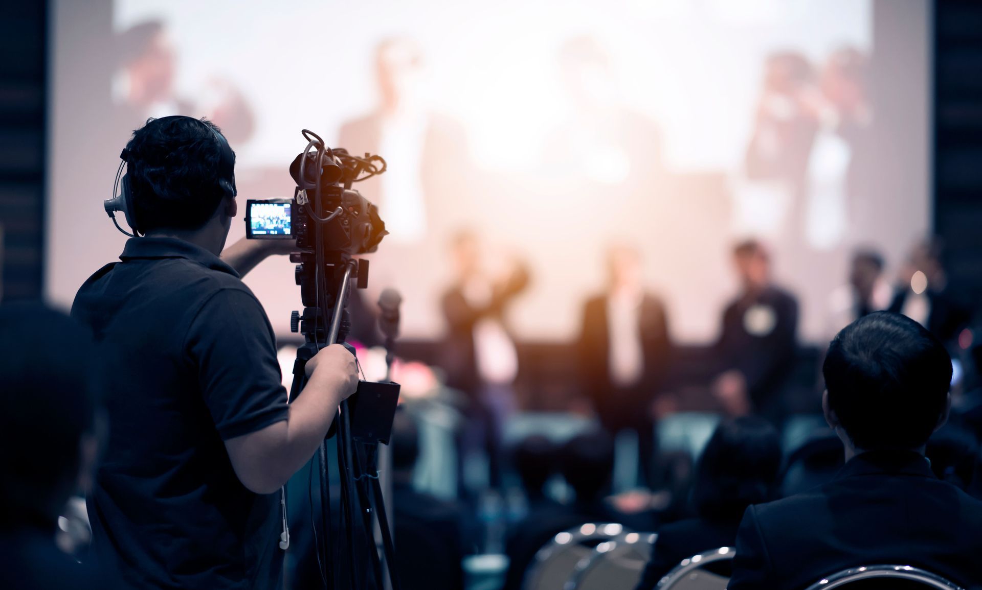 Videographer filming a stage presentation with a camera on a tripod in a dim audience hall