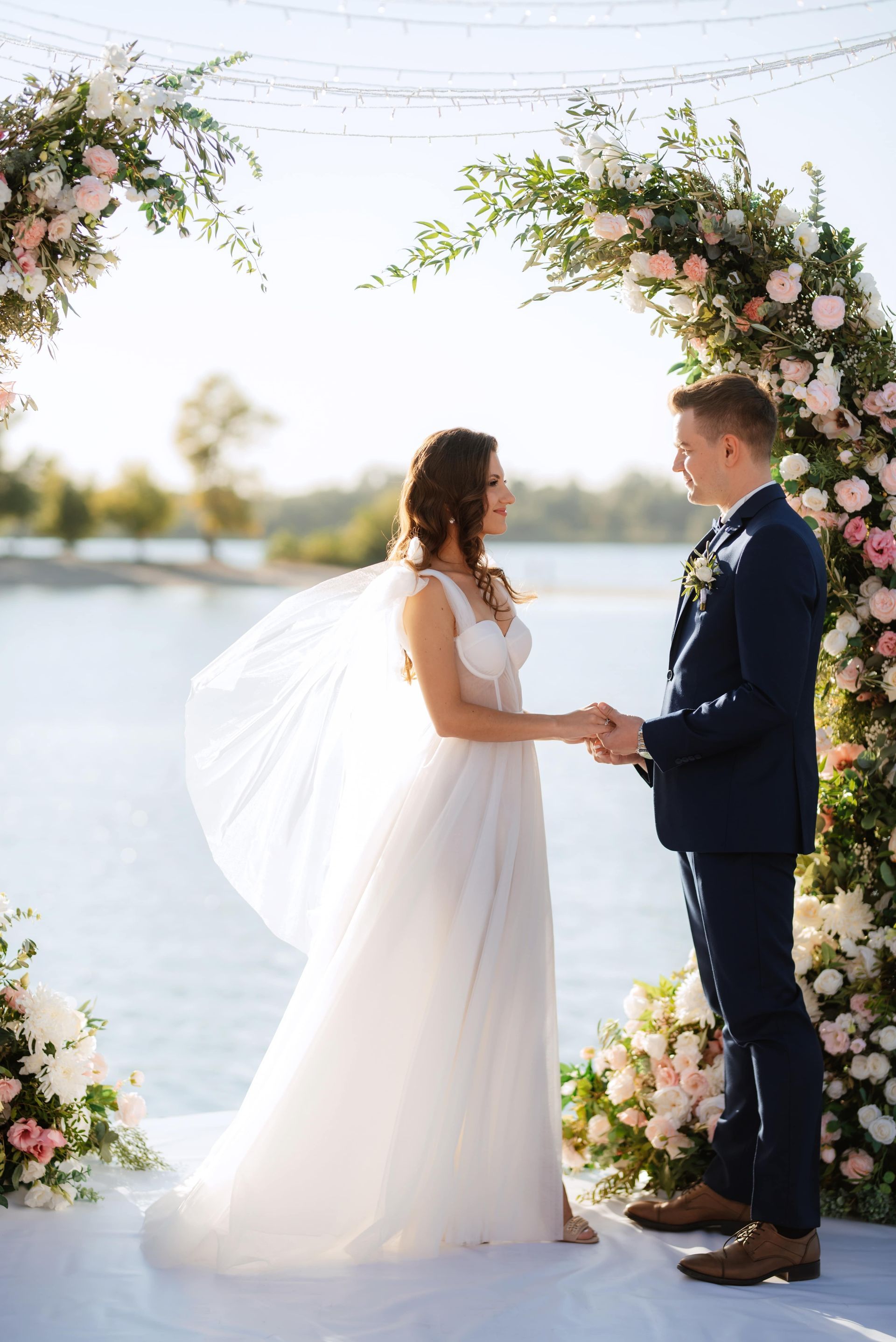 Couple holding hands under floral arches by the water during an outdoor wedding ceremony