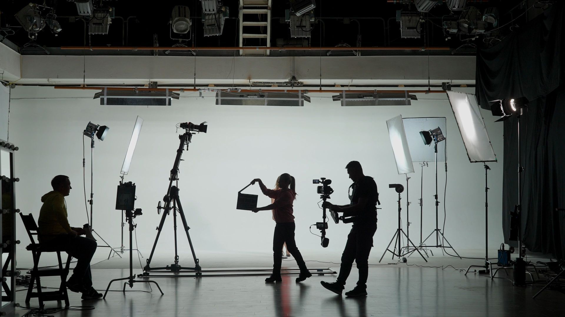 Film studio set with crew silhouetted against a white backdrop, cameras and lights on tripods.