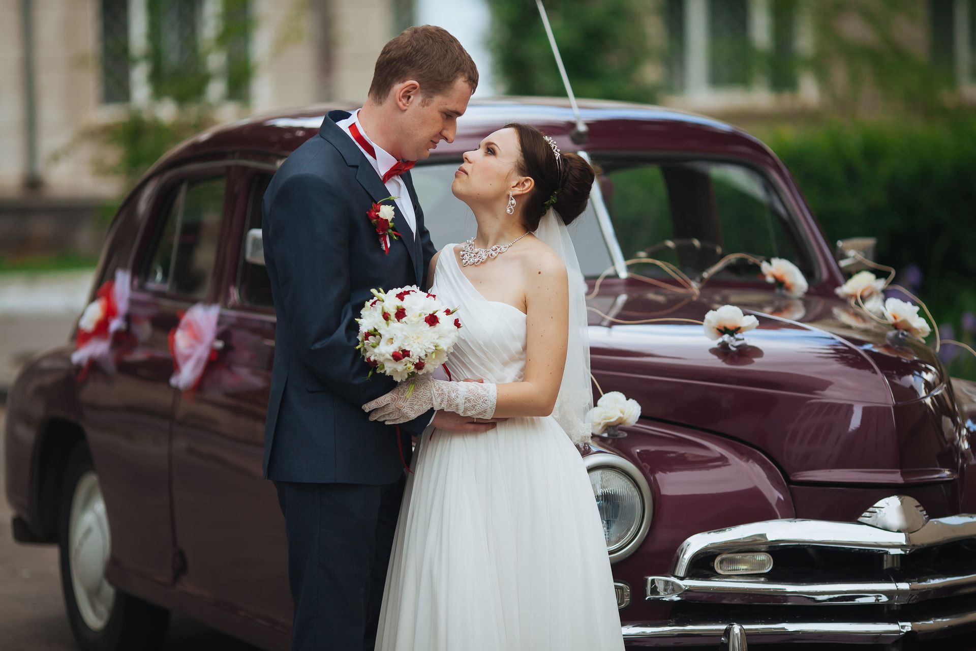 Bride and groom embracing beside a maroon vintage car outdoors