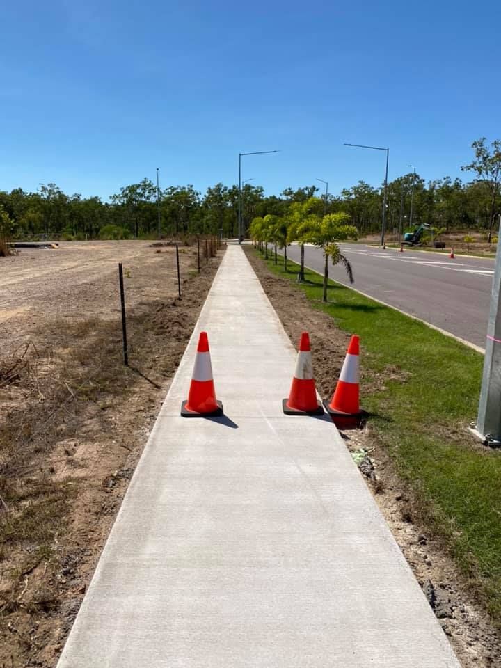 Two traffic cones on a newly paved sidewalk, with a road and trees in the background under a blue sky.