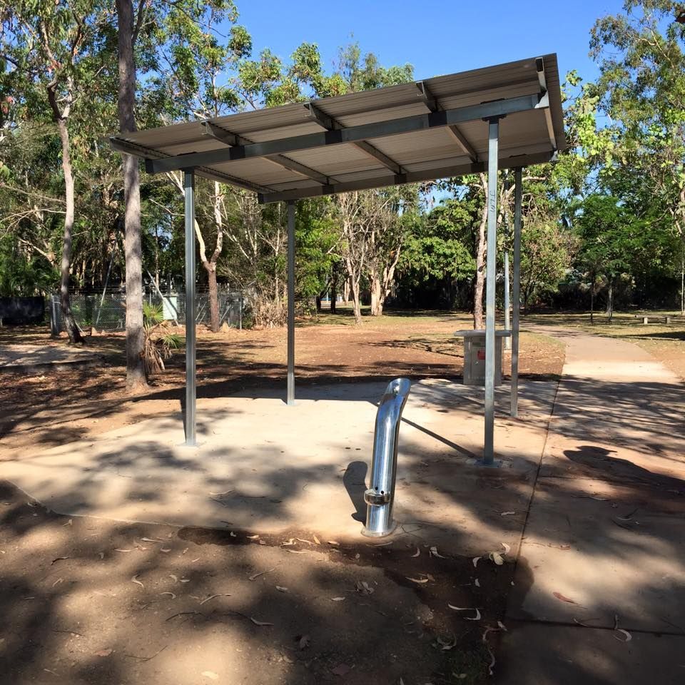 Sheltered water fountain in a park, under a metal roof. Concrete pad, trees in the background, sunny day.