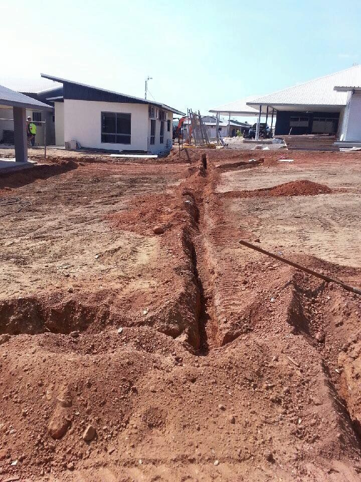 Trenches dug in red dirt at a construction site, near buildings with white walls and blue sky.
