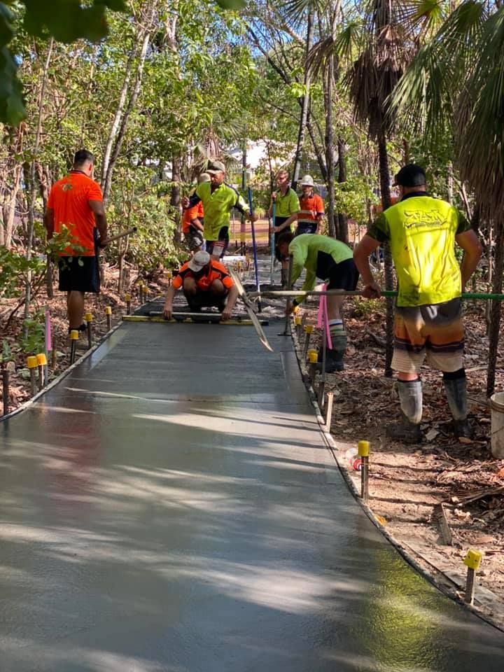 Construction workers pouring concrete on a path in a wooded area. Workers wear neon vests and are using tools.