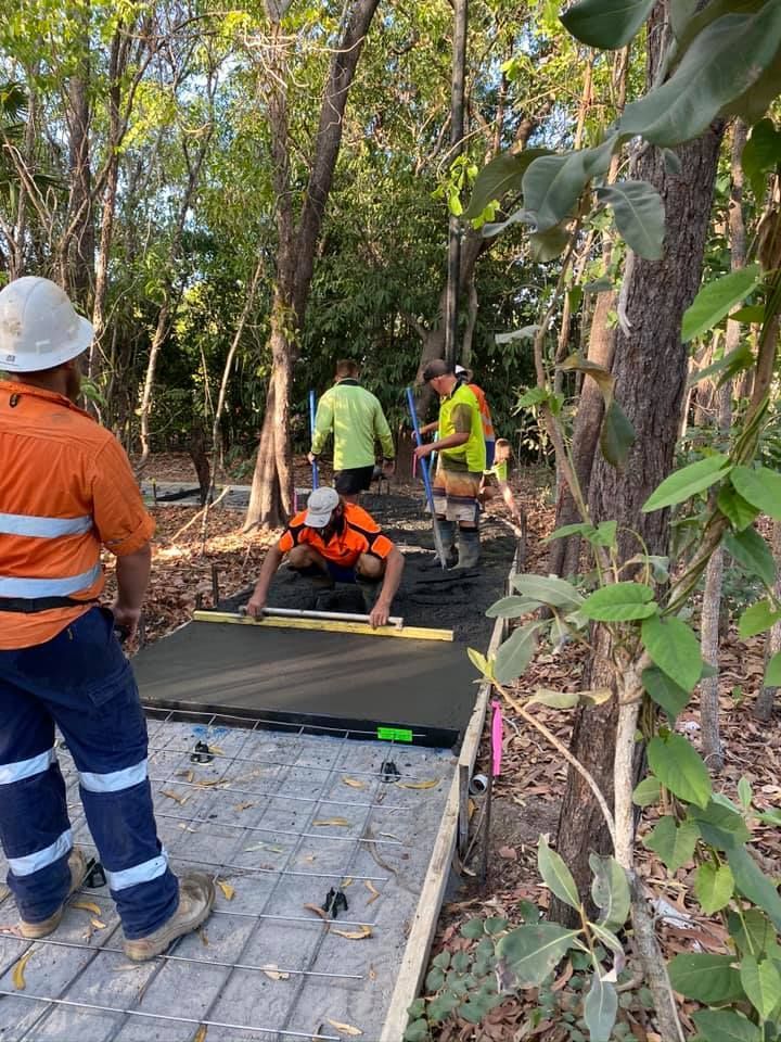 Construction workers leveling concrete pathway in a wooded area; one kneels with a level.