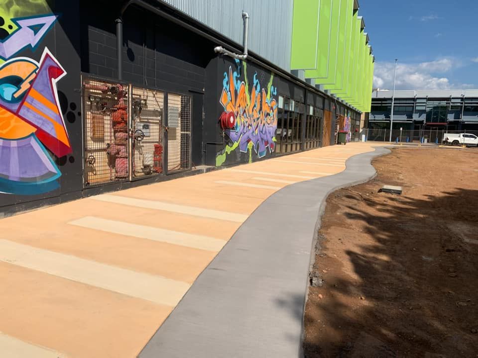 Sidewalk curves along building with graffiti art, beside dirt patch under a blue sky.