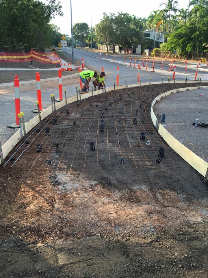 Road construction site: metal grid framework, workers in yellow vests, orange traffic cones, and curved curb.