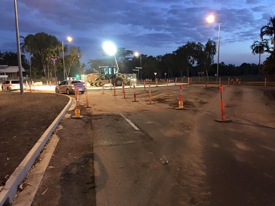 Construction zone at dusk with cones, a grader, cars, and streetlights.