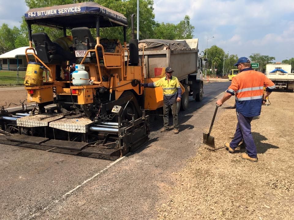 Road paving: Asphalt paver laying fresh asphalt; two workers nearby, one shoveling.