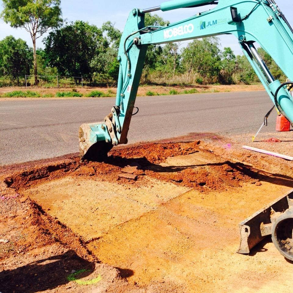 A teal excavator digs into a reddish-brown patch beside a road on a sunny day.