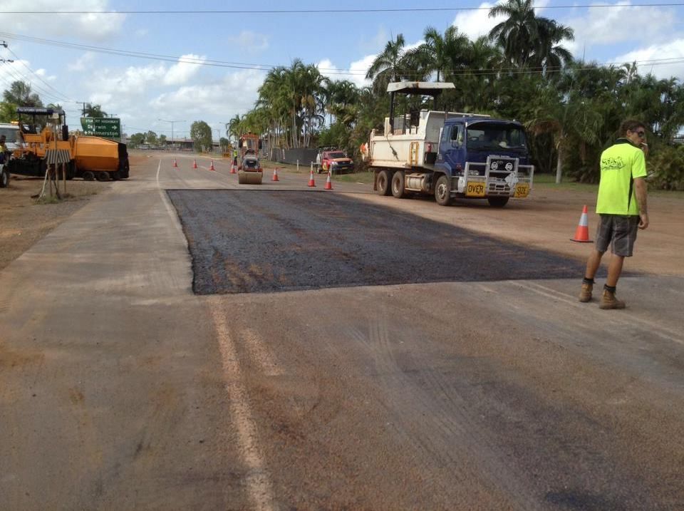Road resurfacing: Dump truck, rollers, workers, and orange cones on a street under a blue sky.