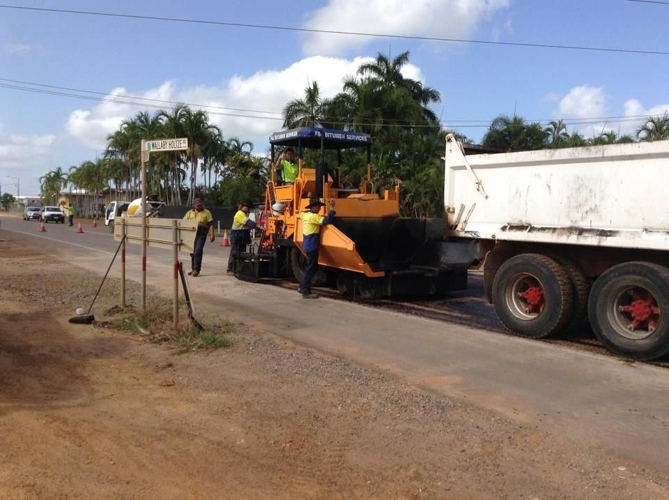 Road paving in progress; asphalt machine laying new road surface; dump truck delivers material; workers in vests.