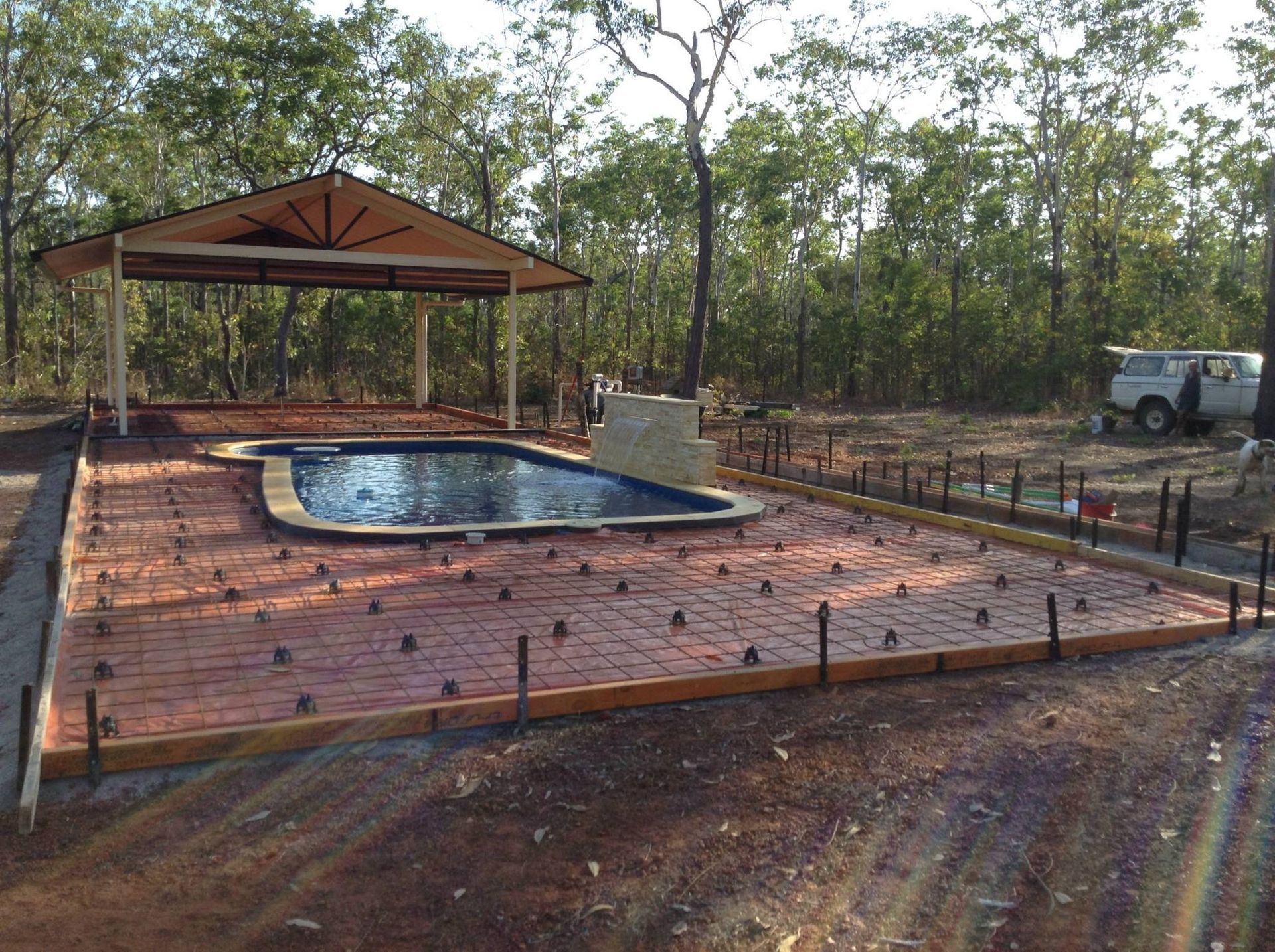 Swimming pool with a pavilion, brick patio, surrounded by trees and a vehicle in the background.