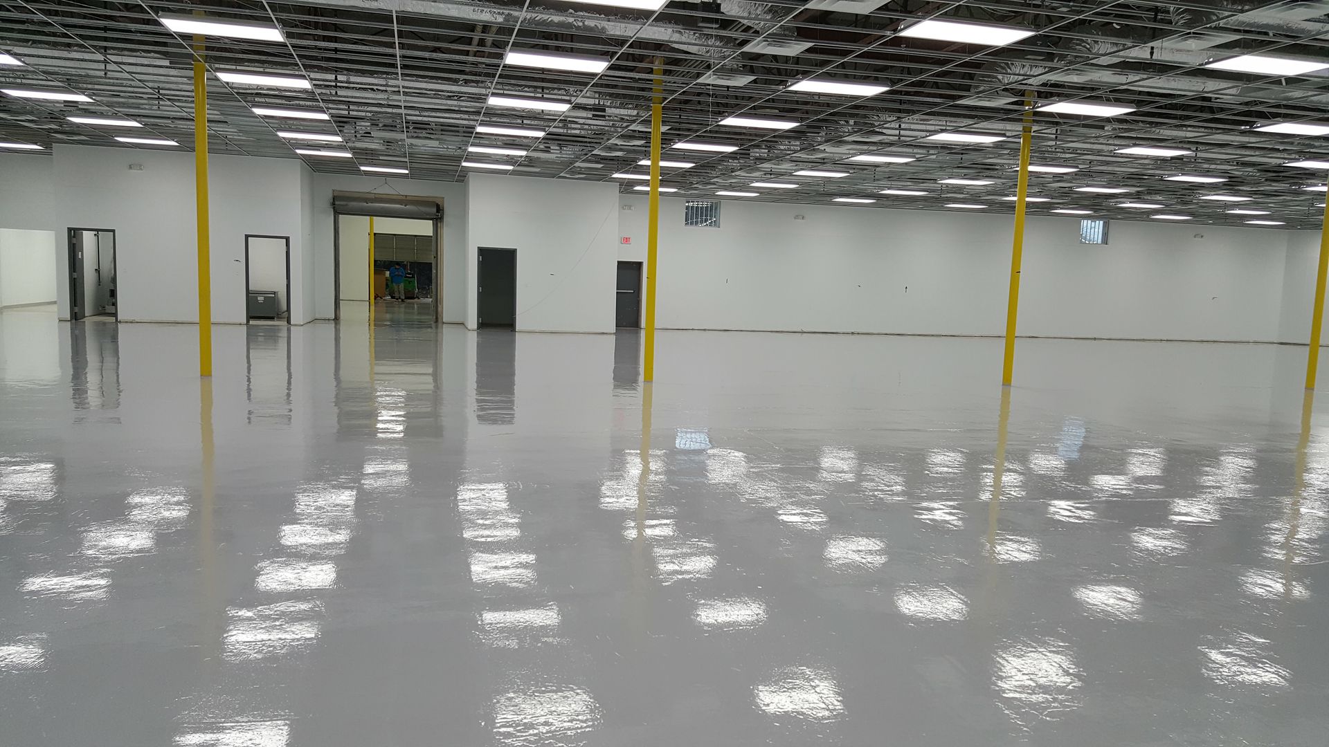 A spacious, empty commercial room with a high, exposed ceiling, yellow support pillars, and a reflective gray epoxy floor.
