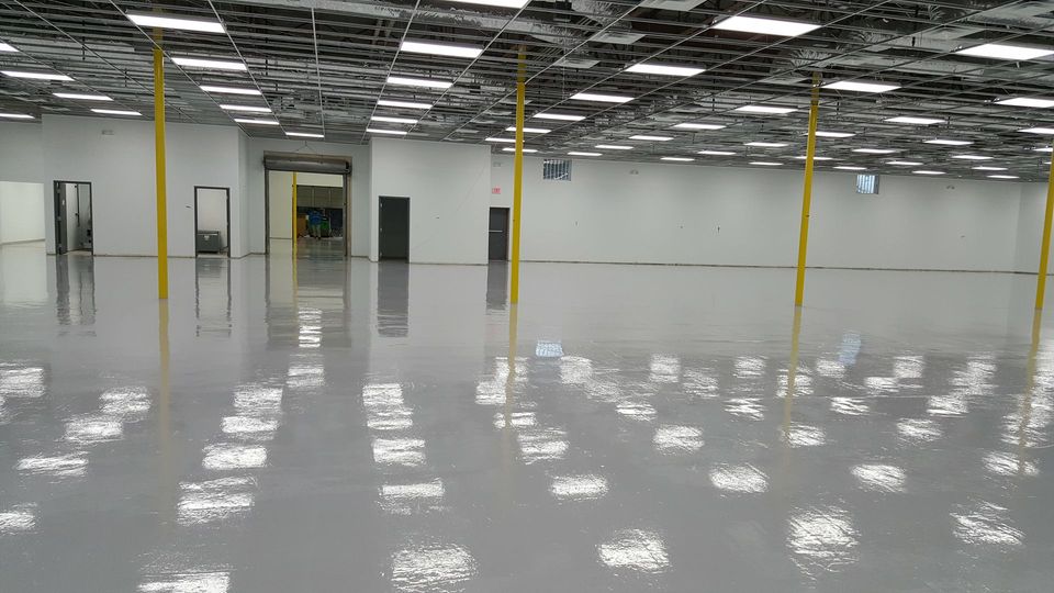 A large, empty warehouse space with shiny, gray epoxy floors and bright, reflective ceiling lights.