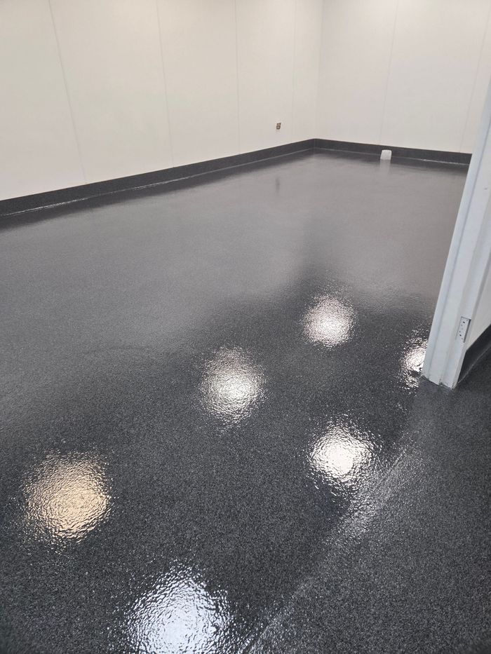 An empty room with light grey walls, dark baseboards, and a glossy, speckled grey epoxy floor reflecting ceiling lights.