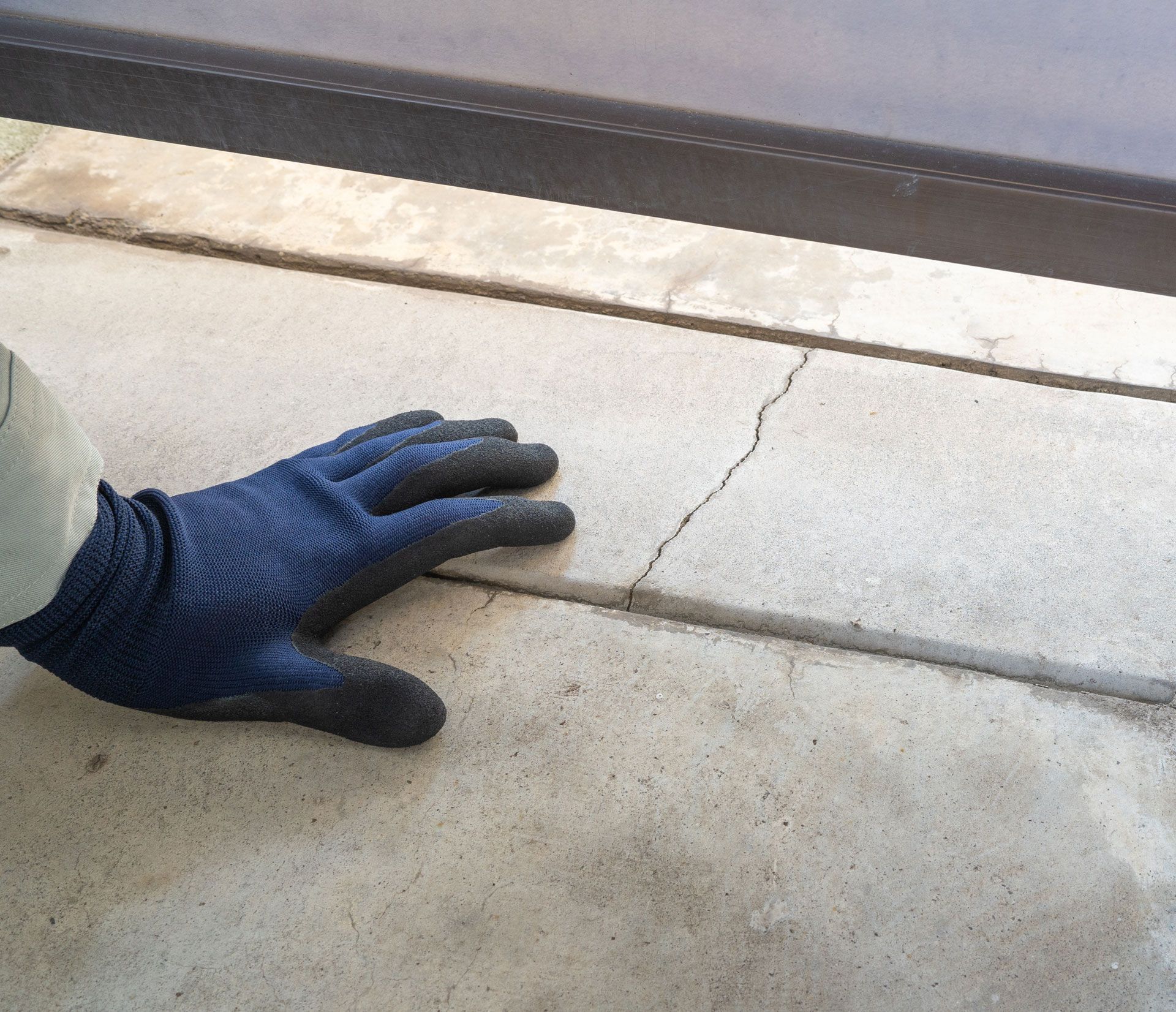 A person wearing a dark blue work glove points to a crack in a concrete patio slab near the base of a dark metal fence.