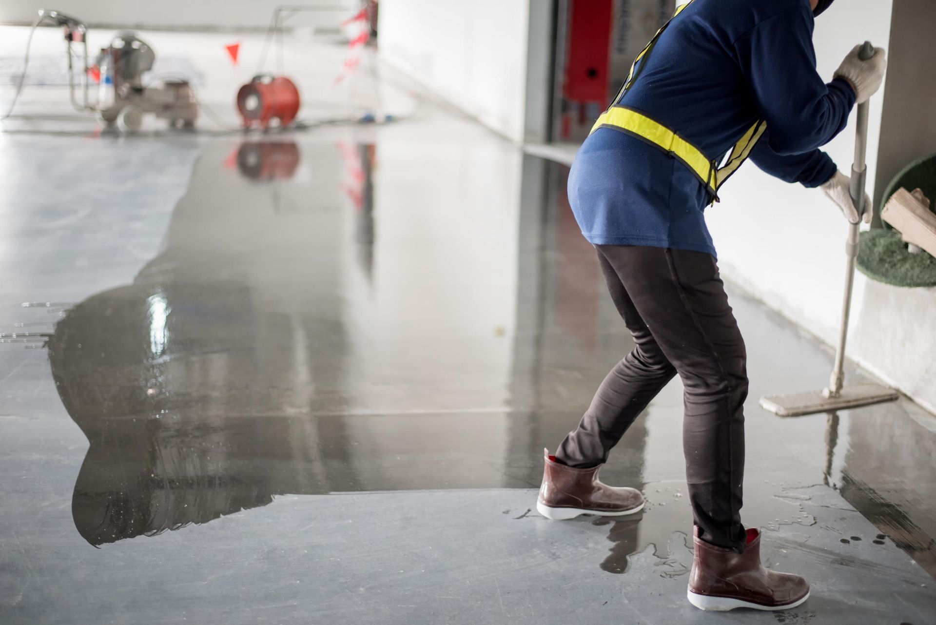 A worker in safety gear applies a glossy, wet coating to a concrete floor inside a building.