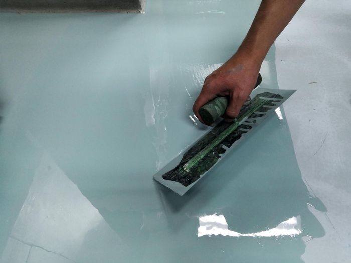 A hand uses a flat trowel to smooth light blue epoxy resin across a floor.