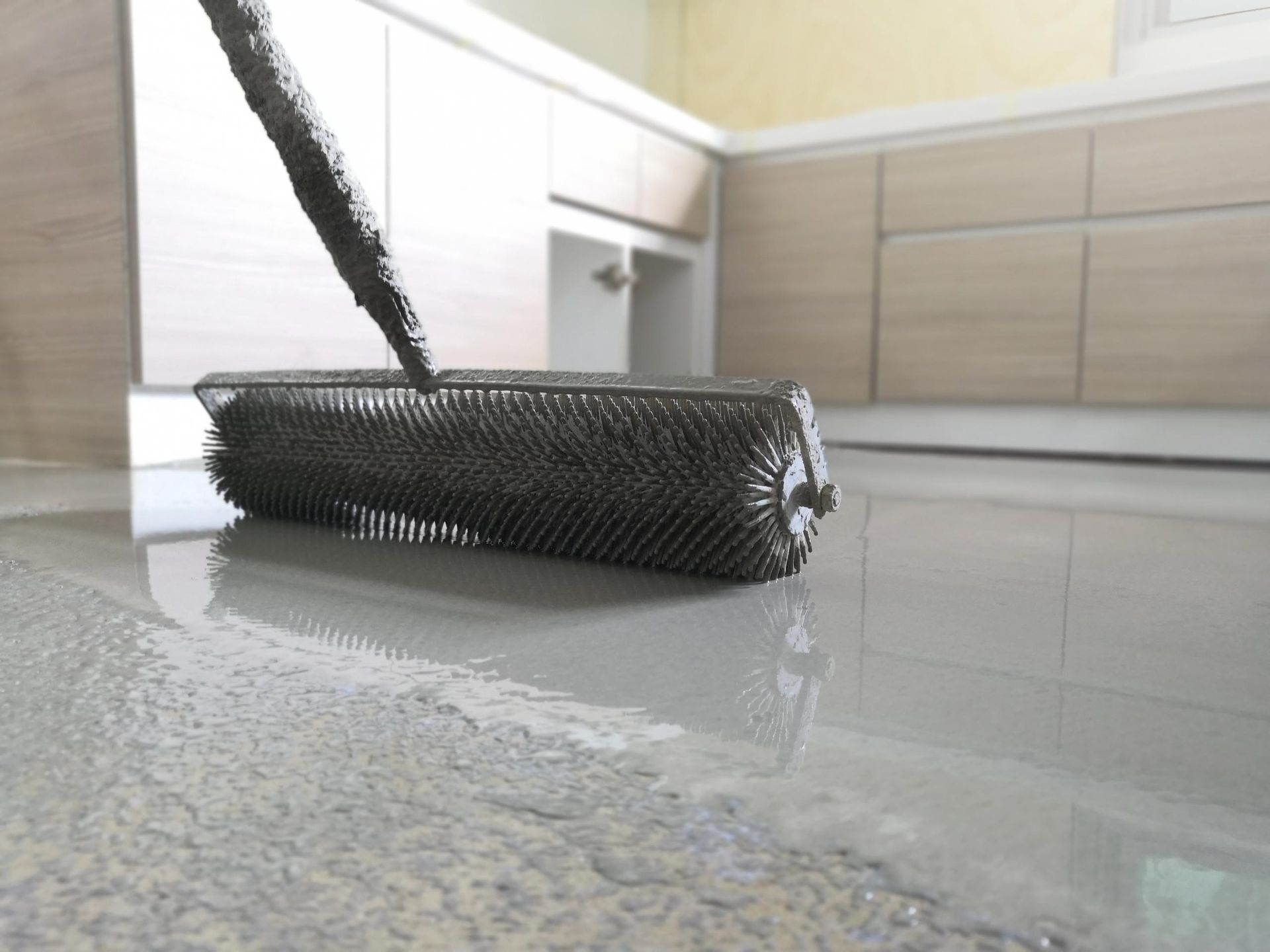 A spike roller spreads grey epoxy coating across a kitchen floor to remove air bubbles.