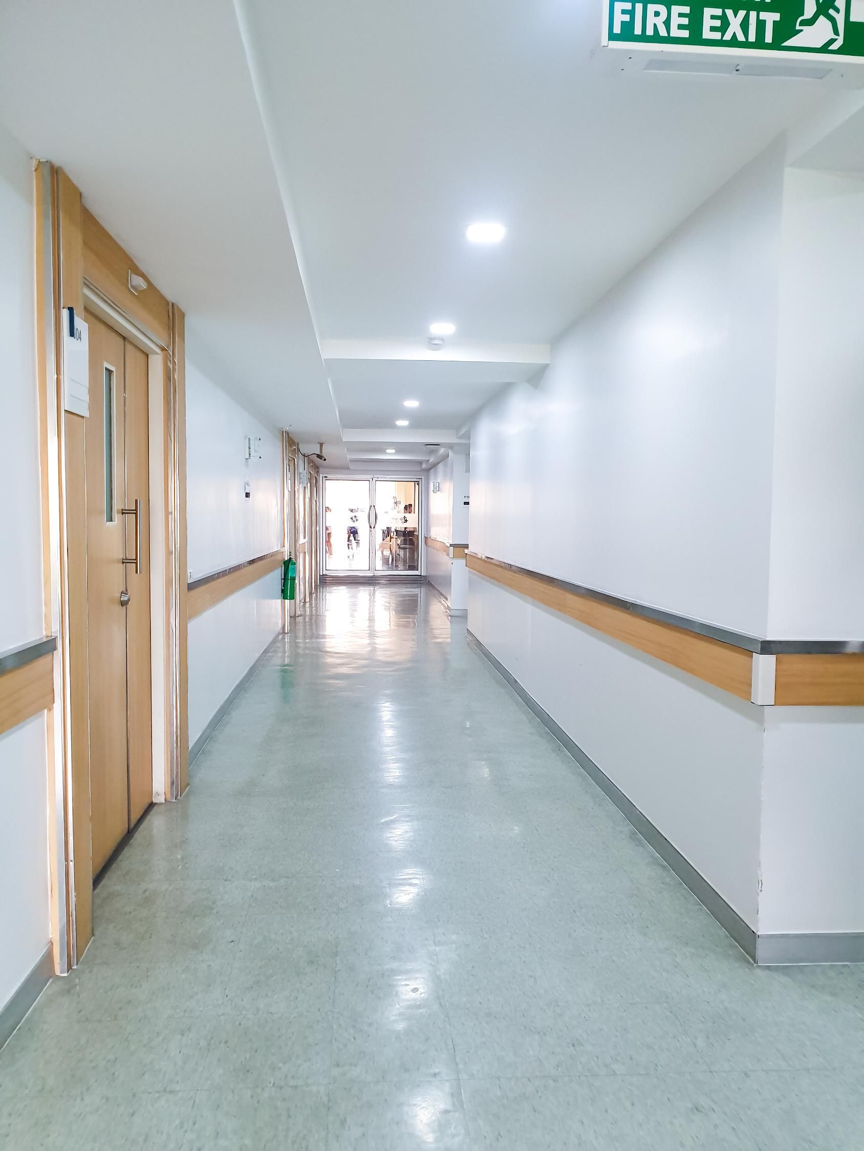 A well-lit hospital hallway with light-colored walls, wood-trimmed railings, and a green fire exit sign at the end.