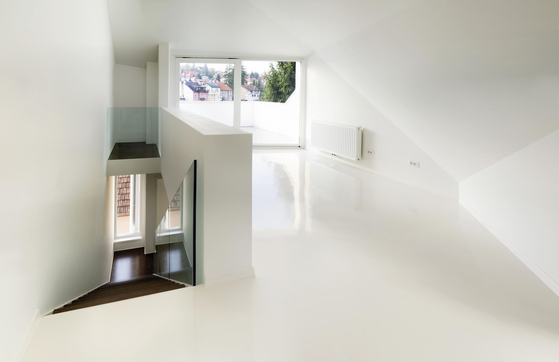 A bright, minimalist attic room with white walls, a glossy white floor, and a glass-railed staircase leading downstairs.