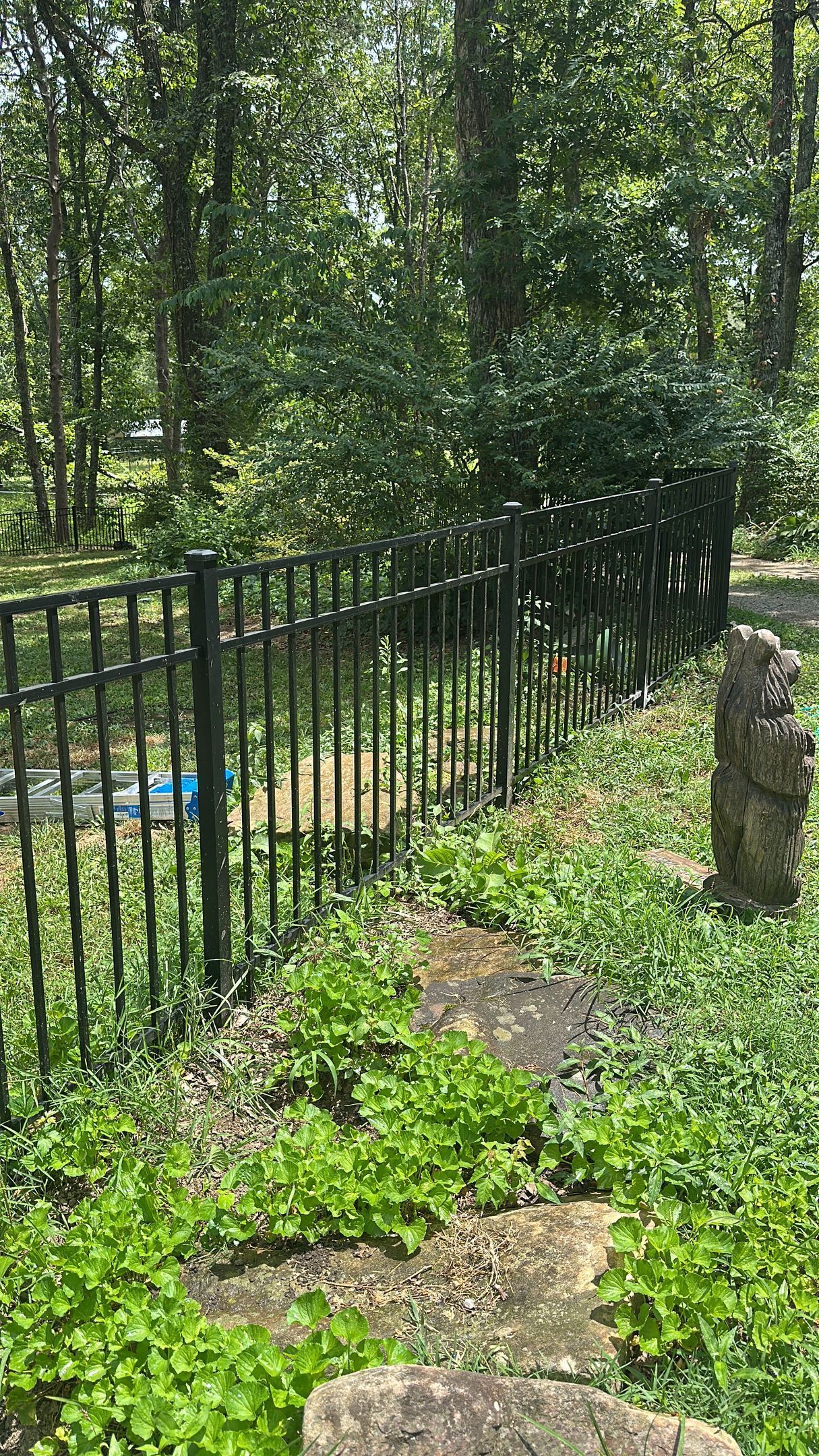 Stone path beside black metal fence in a wooded yard, with a stone post and green ground cover.