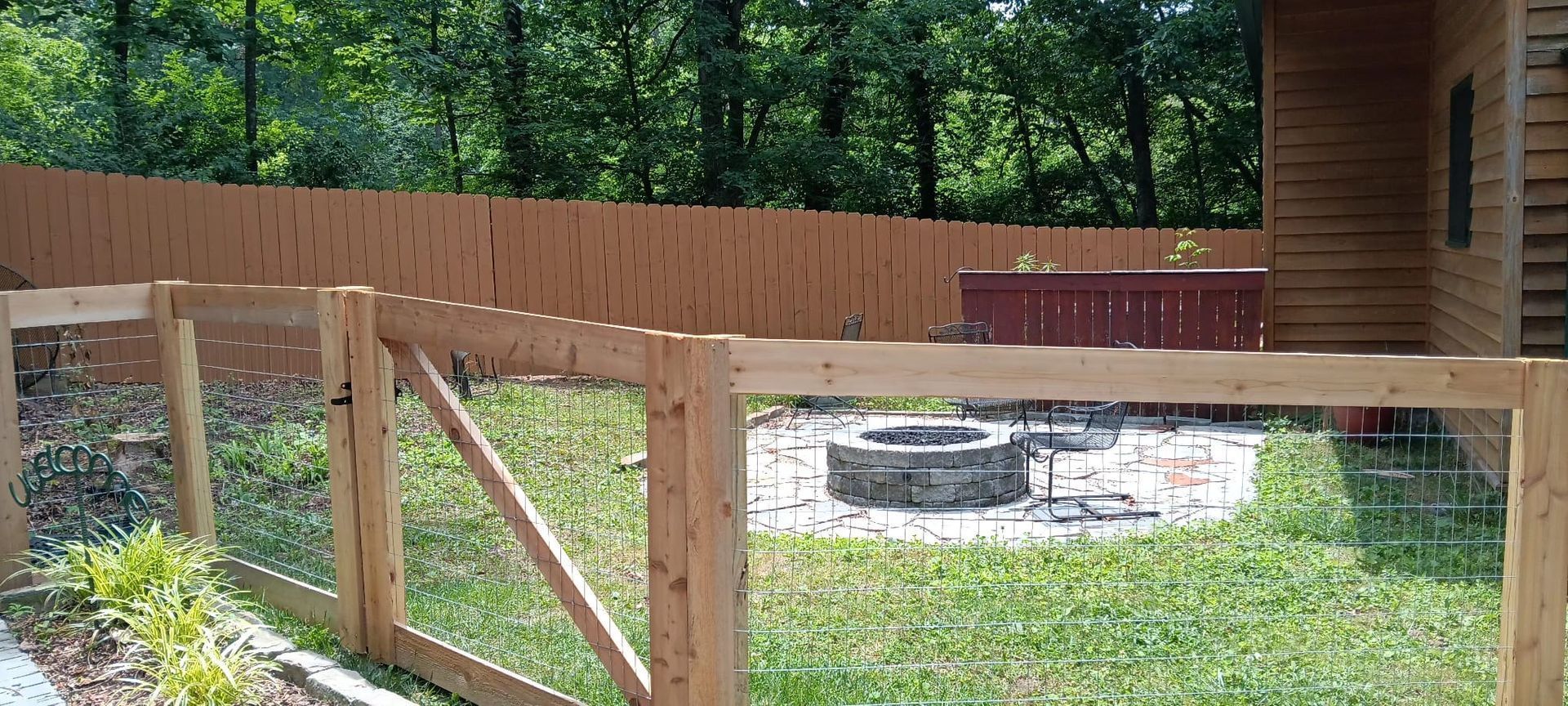 Wooden fence around a grassy yard with a stone fire pit beside a log cabin and trees