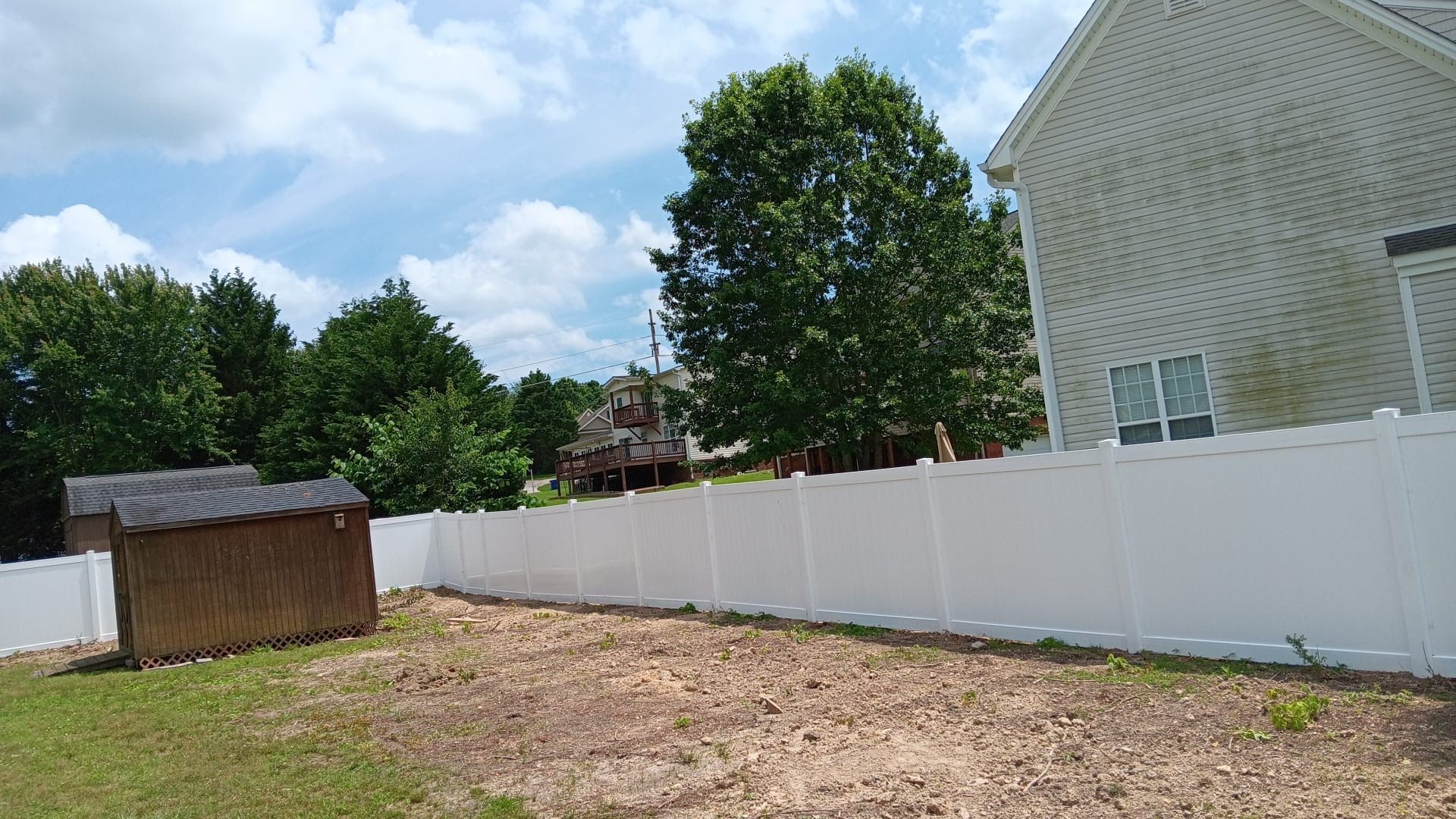 Backyard with white fence, small shed, trees, and a house under a blue sky
