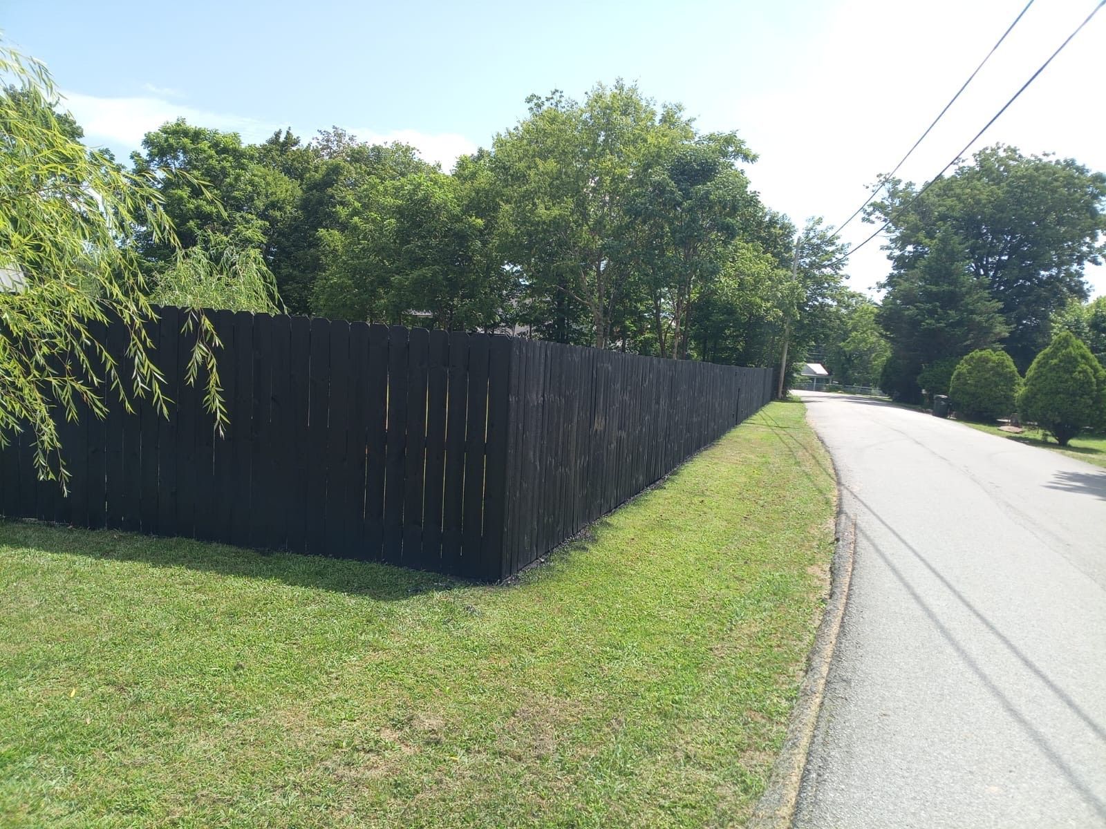 Black wooden fence along a grassy roadside under bright sunlight, with trees and a paved street.
