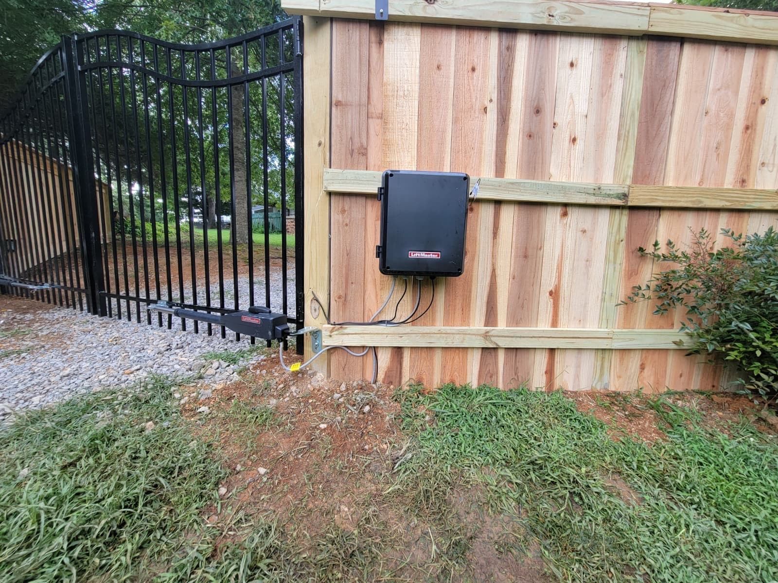 Black metal gate beside a wooden fence with a mounted black box and gravel driveway