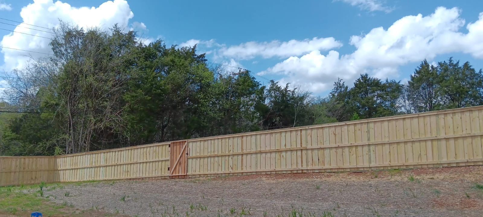 A long wooden fence with a central gate under a blue sky, with trees in the background.