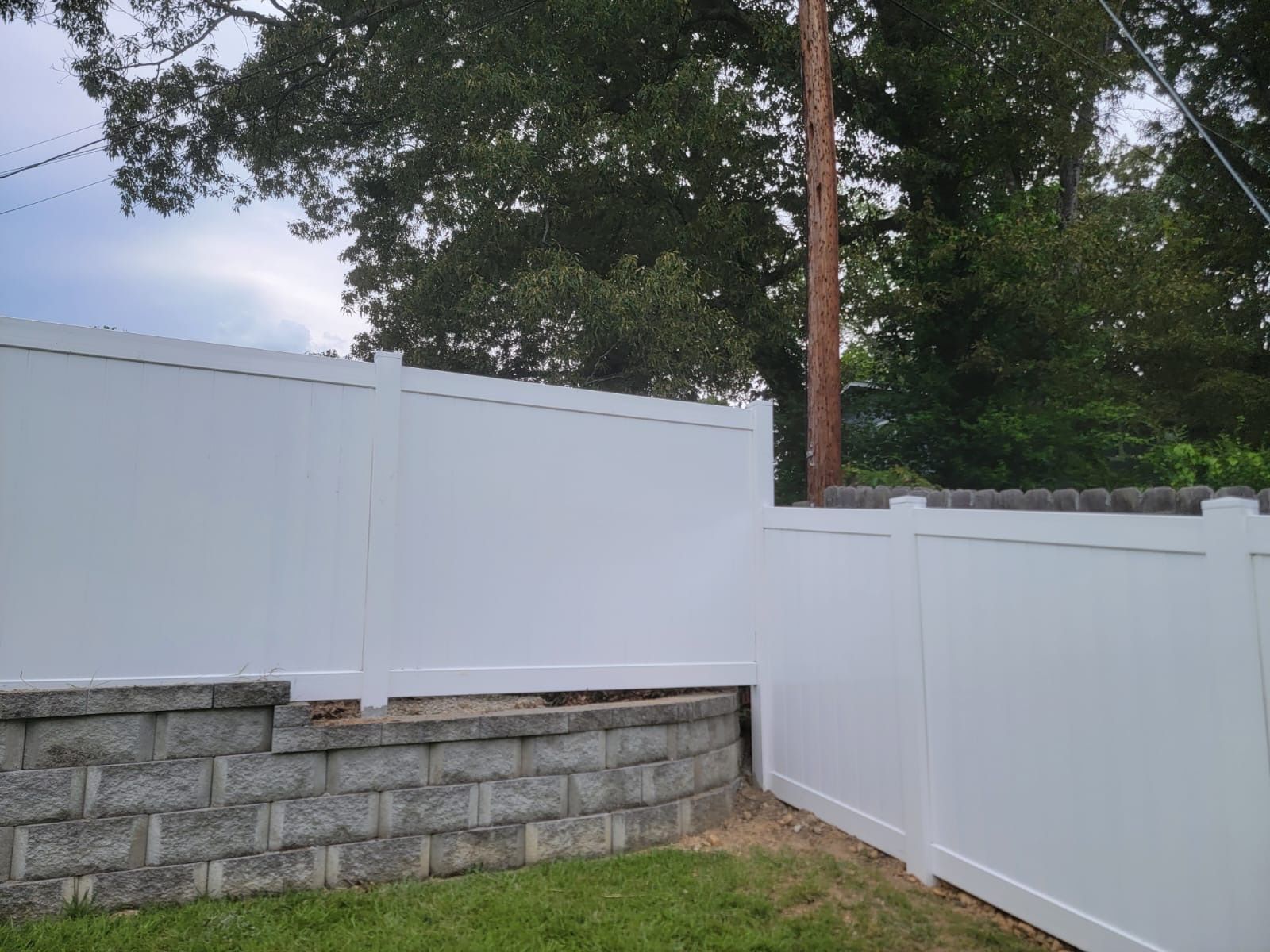 White vinyl fence along a stone retaining wall beside a tree-lined yard under a cloudy sky