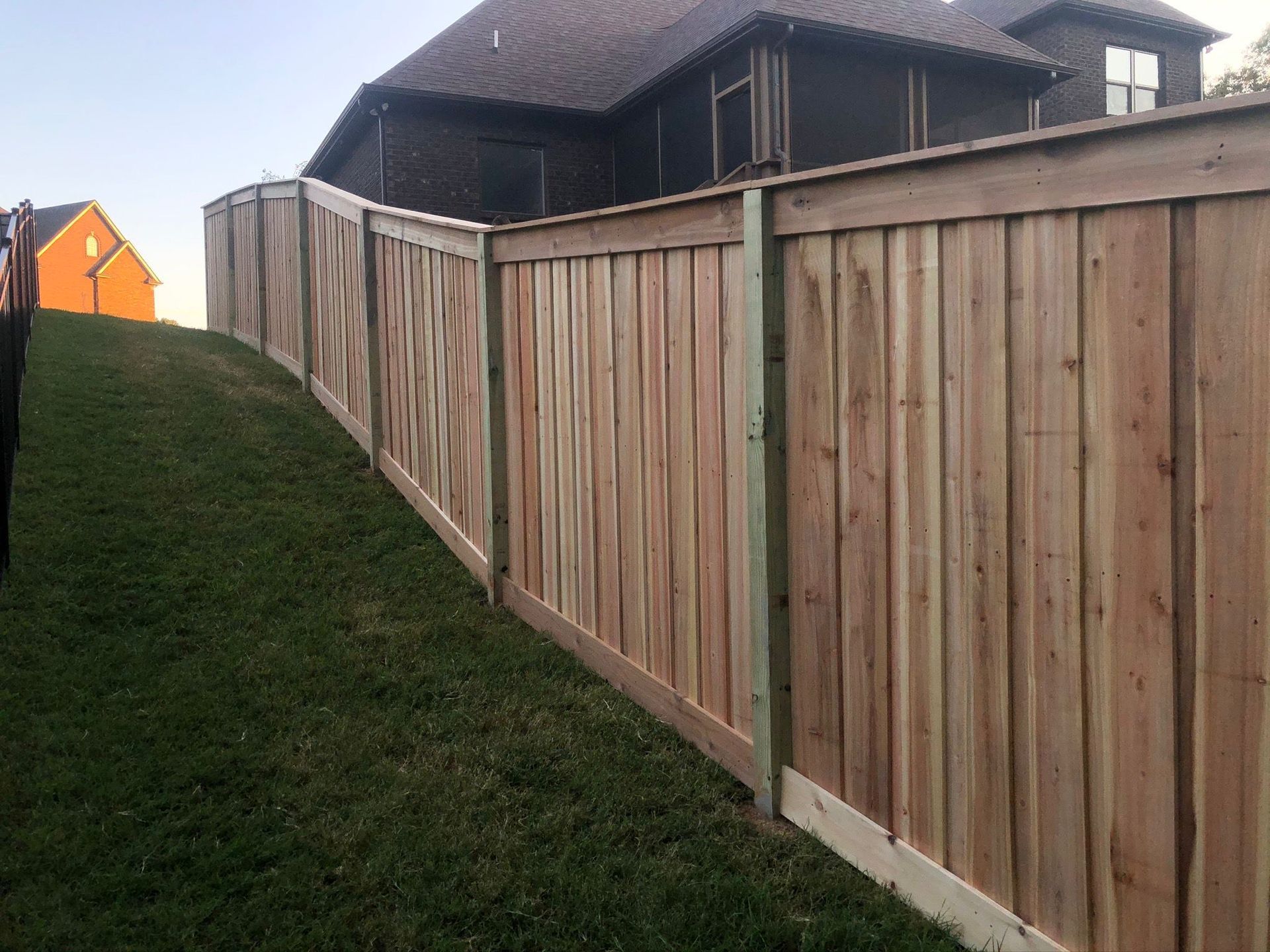 Wooden fence along a grassy slope beside houses, with an orange house in the distance.