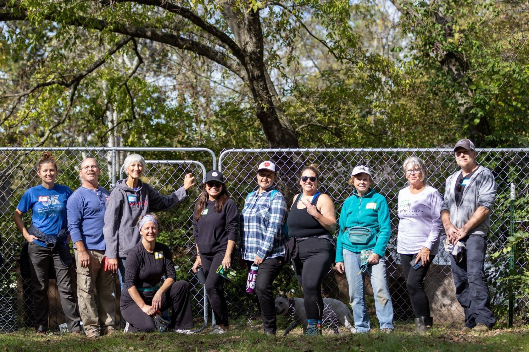 Group of people posing outdoors in a sunny park with trees and a chain-link fence behind them
