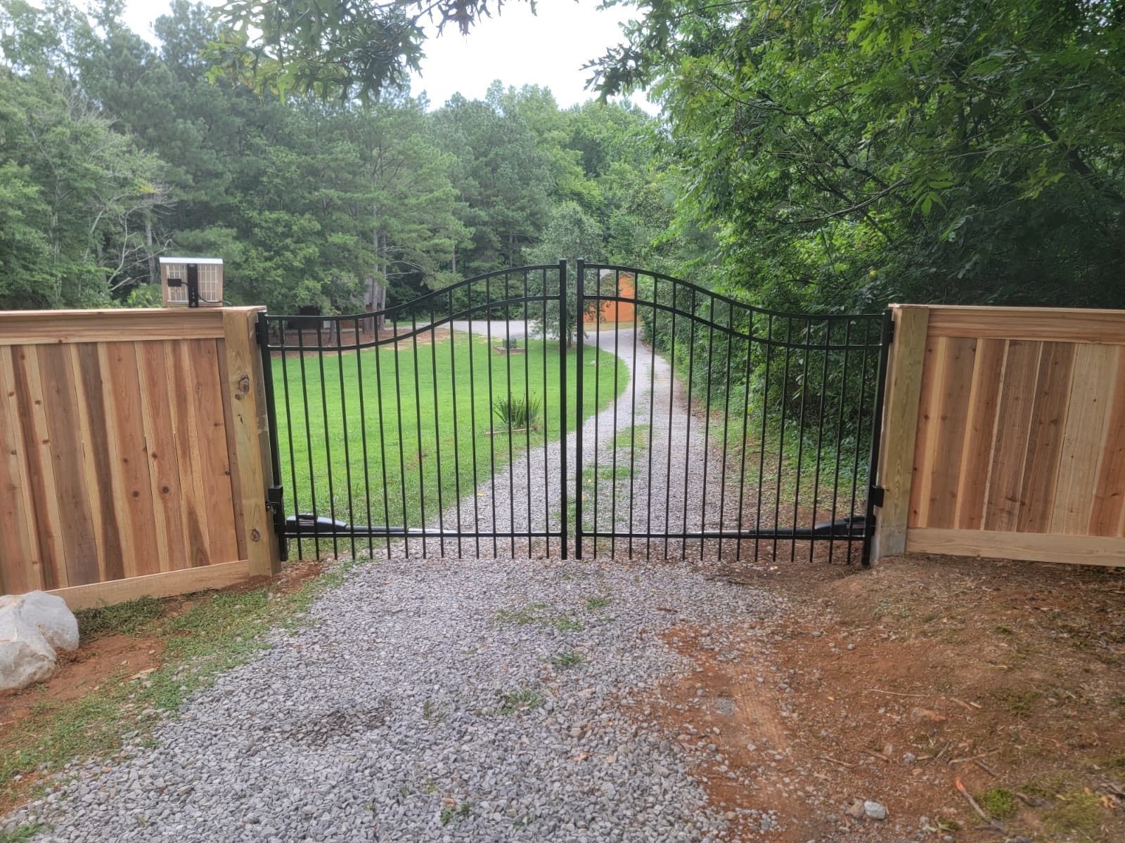 Gravel driveway leading to a black metal gate, framed by wooden fence panels and green trees.