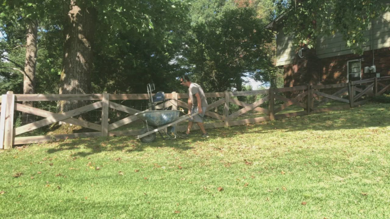 Person working on a wooden fence in a grassy yard with trees and a house in the background