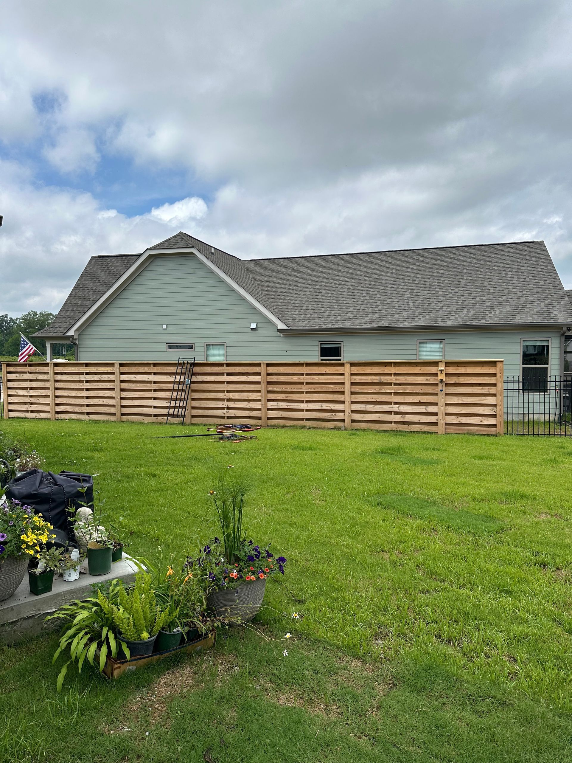 Backyard with green lawn, wooden fence, flower bed, and a gray house under cloudy skies