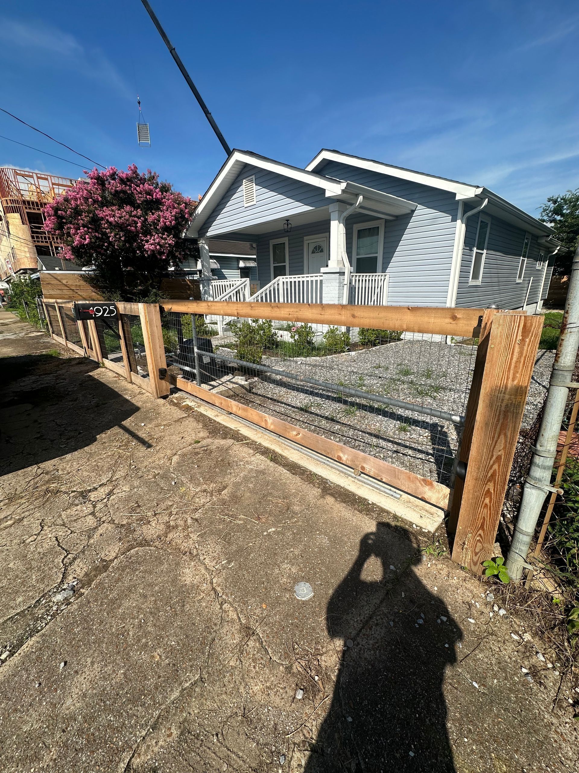 White house with porch behind a wooden gate and gravel driveway under a blue sky.