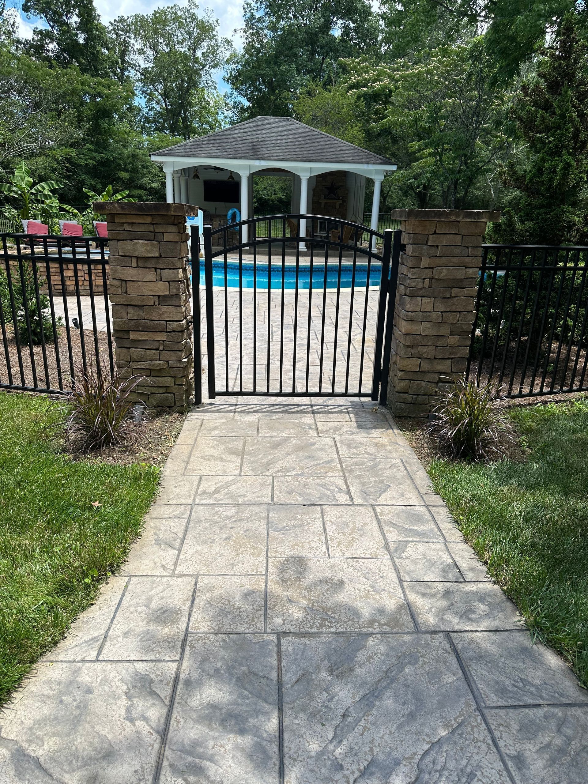 Stone walkway to a gated backyard pool with a white gazebo and trees behind it
