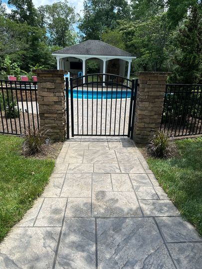 Stone walkway to a fenced backyard pool with a white gazebo and trees behind it