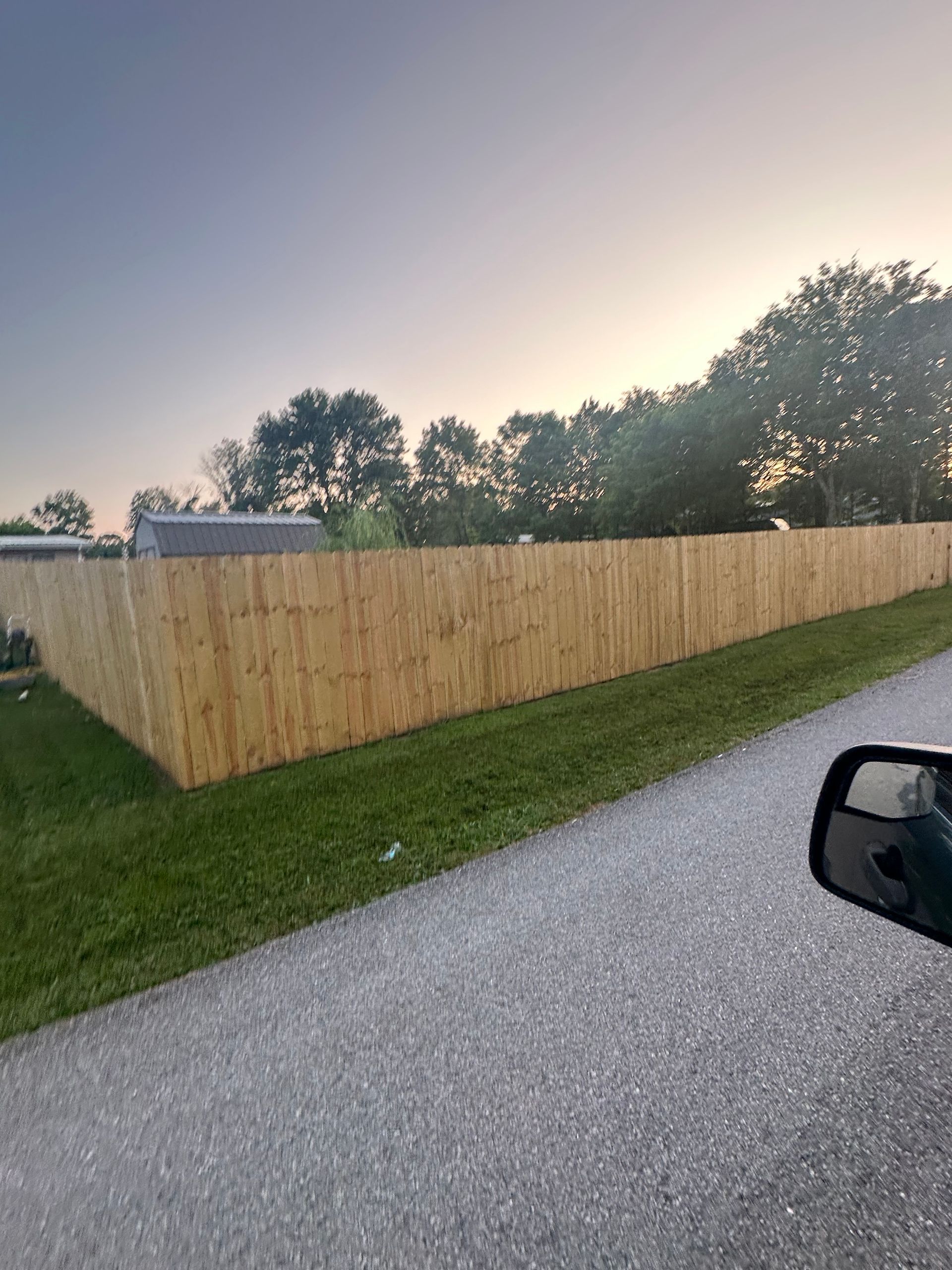 Wooden privacy fence along a gravel drive at dusk, with trees and a parked vehicle visible.