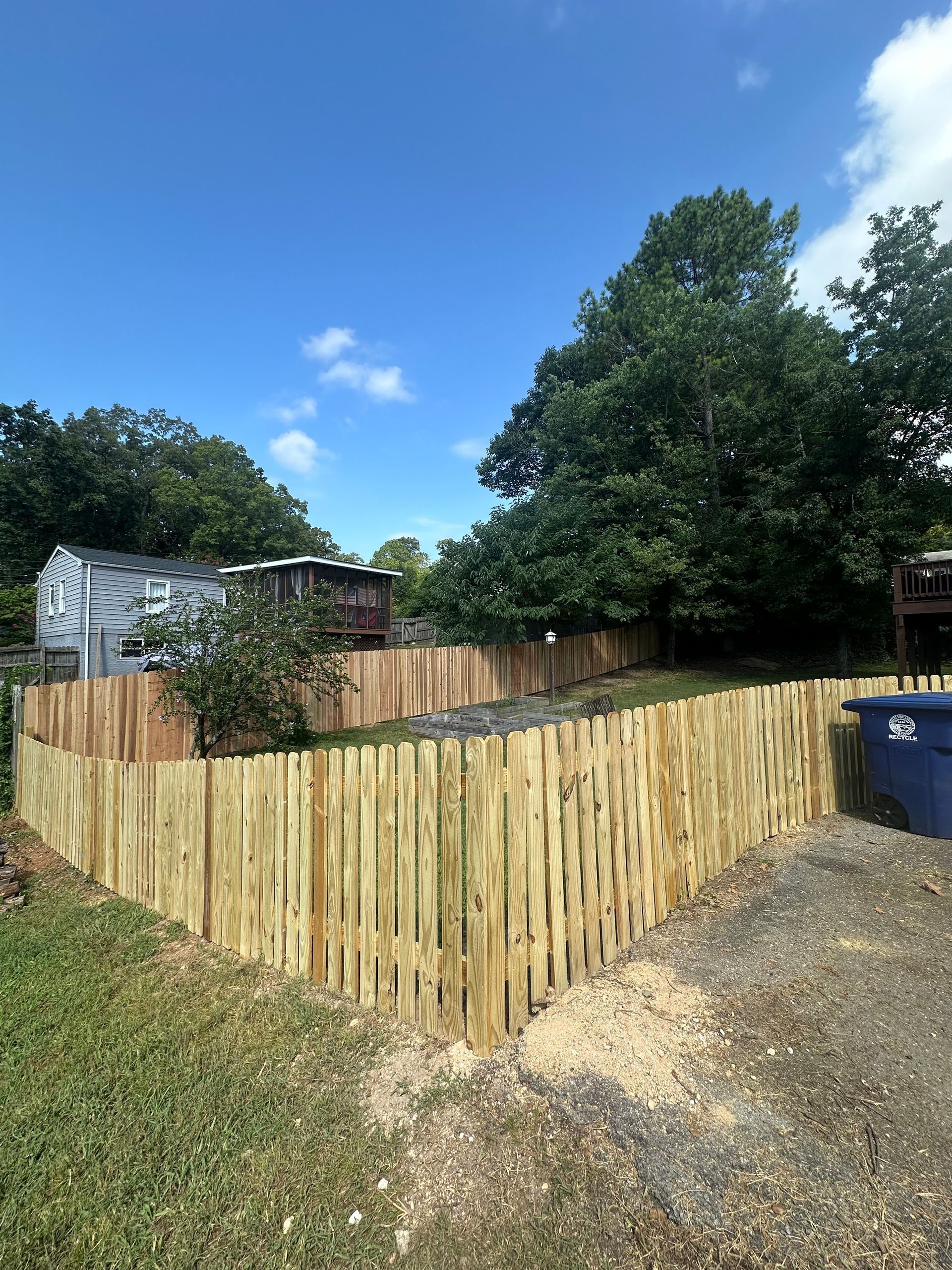 Wooden privacy fence around a yard, with trees, gravel driveway, and blue sky.